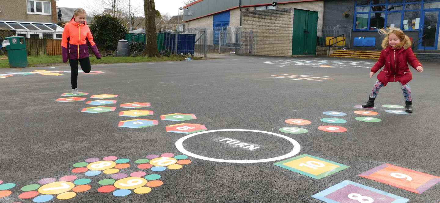 Two girls hopping along numbered hexagon and dot trail markings either side of a white TURN circle on a school playground