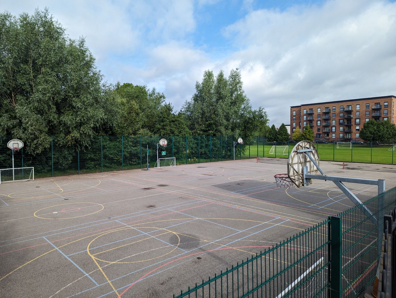 Fenced multi-use games area with overlapping yellow, blue and red court markings, basketball hoops and small football goals