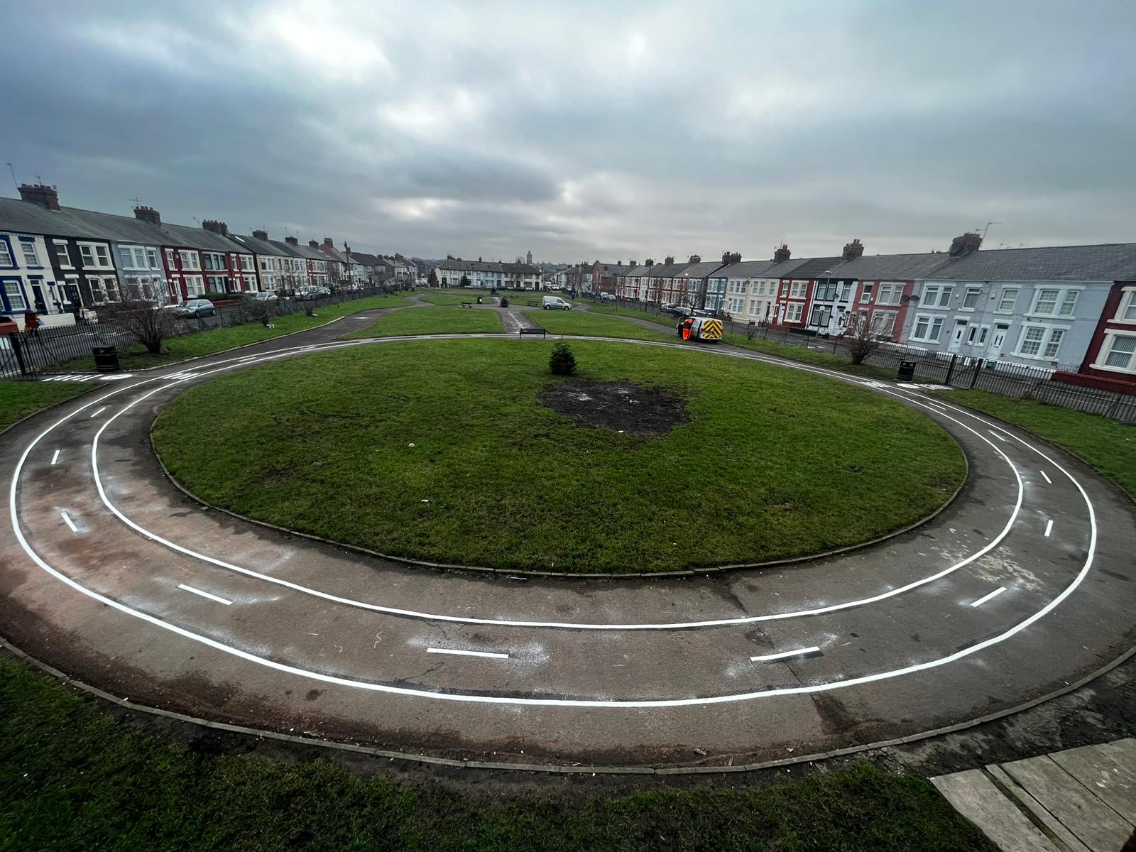 White road track markings with dashed centre lines on a circular path around a park lawn, with terraced houses on both sides