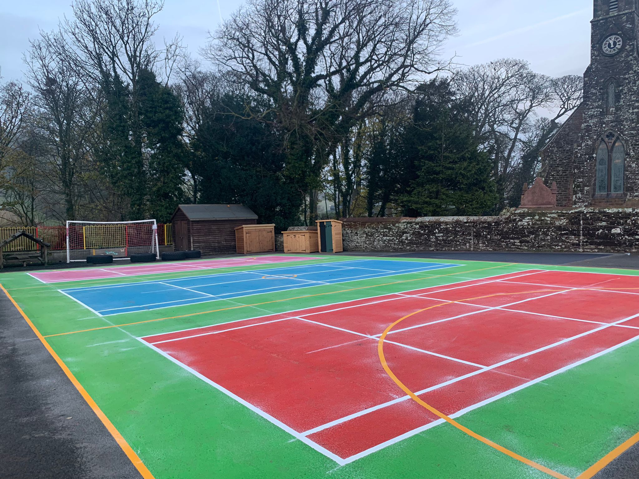 Red, blue and pink tennis courts with orange netball line markings on a green colour-coated playground beside a stone church