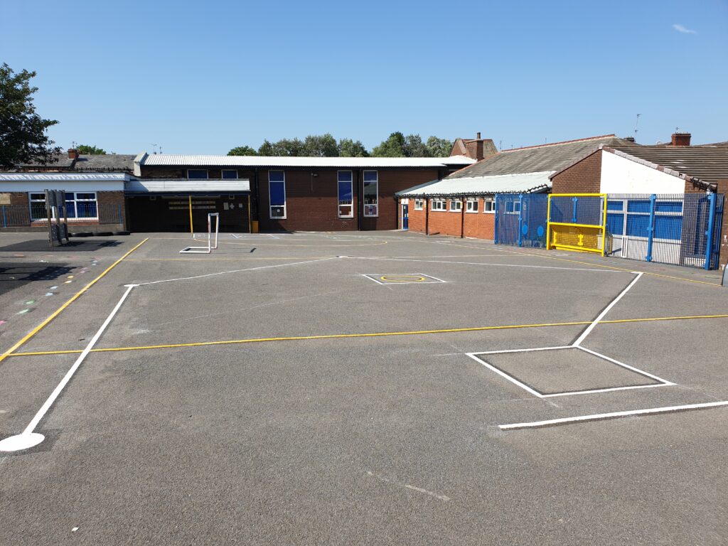 Sunny school playground with new white and yellow game line markings, a small goal and a blue fenced ball court behind