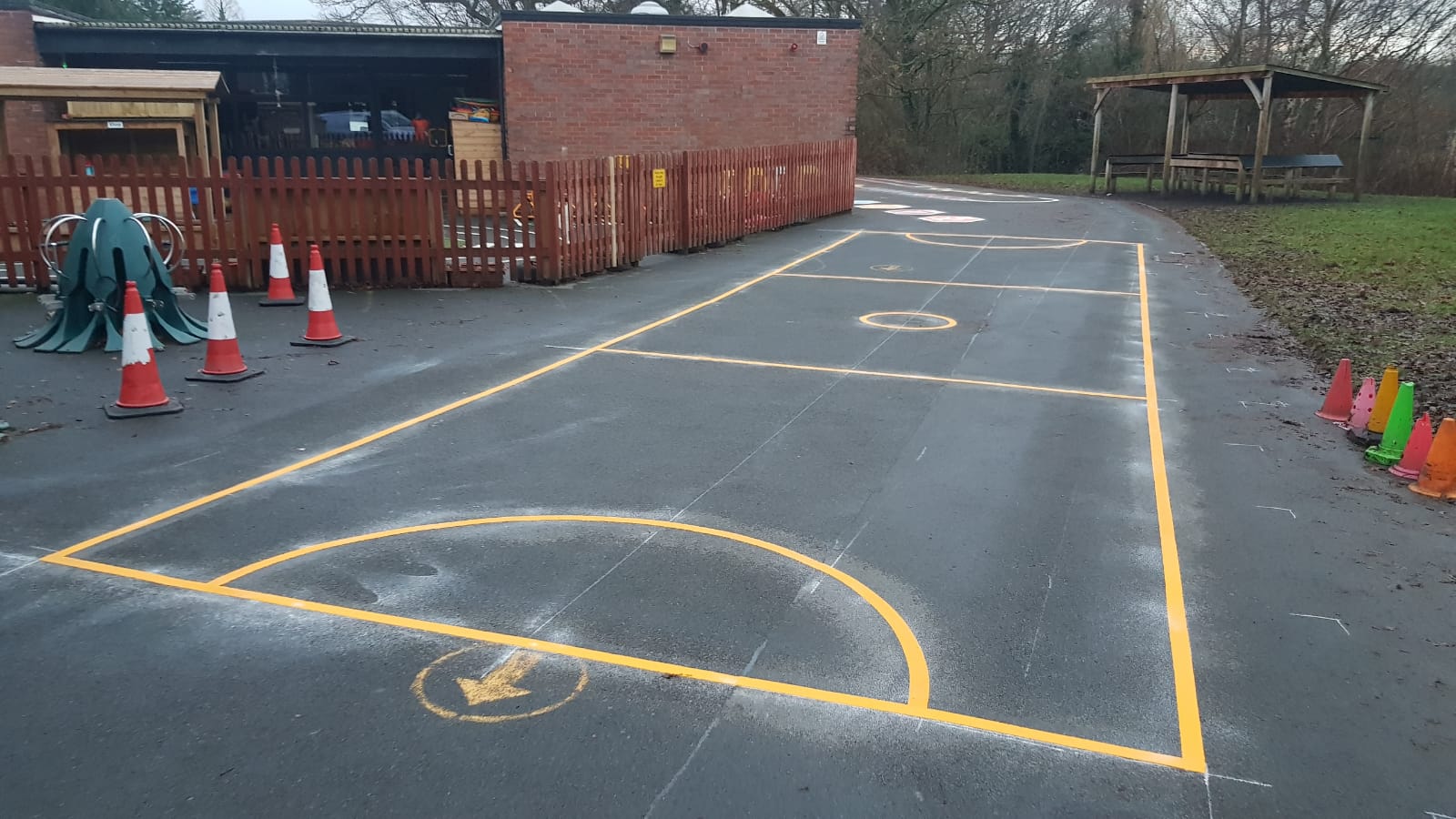 Freshly laid yellow netball court markings on a school playground, with cones, a picket-fenced play area and a wooden shelter