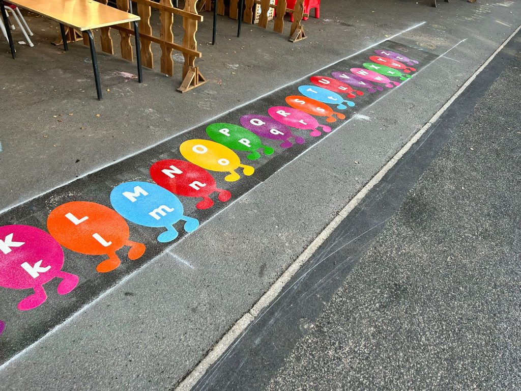 Thermoplastic alphabet trail of colourful oval characters with feet, each showing upper and lower case letters, by outdoor tables