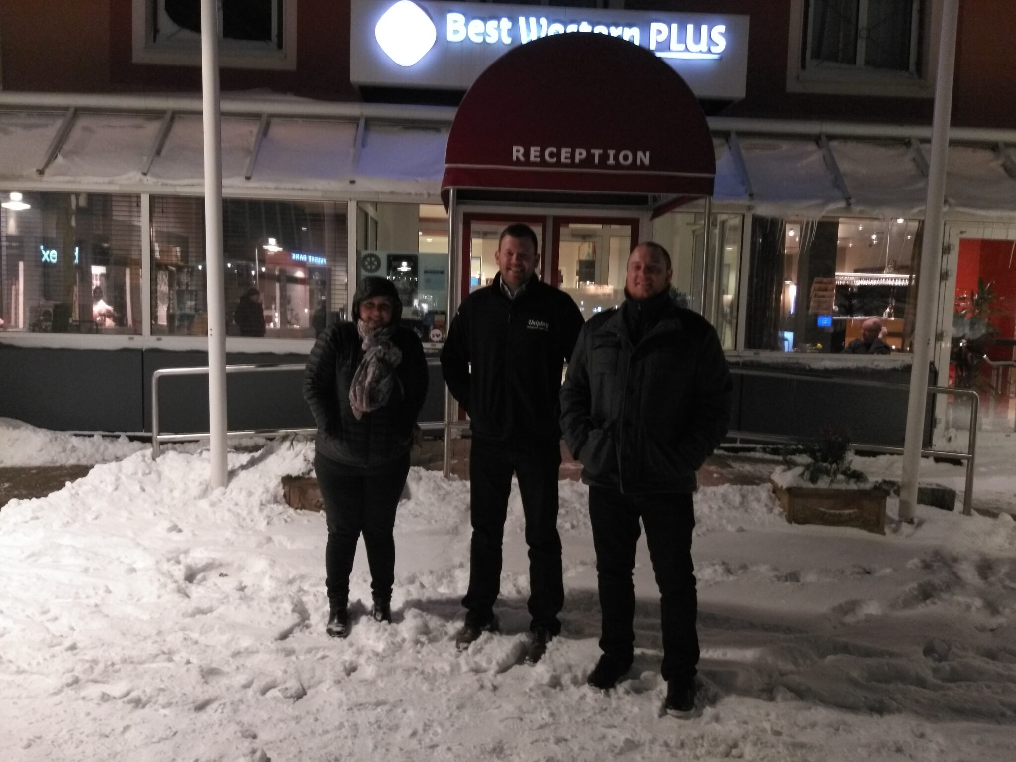 Three people in winter coats, one wearing a Uniplay jacket, standing in snow outside a lit hotel reception at night