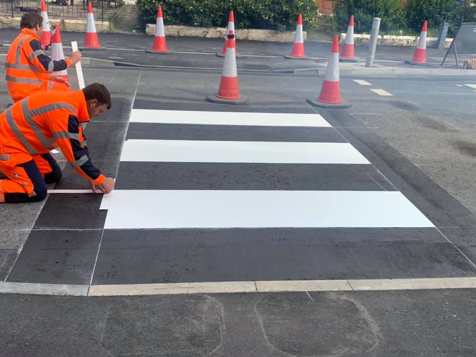 Two installers in orange hi-vis laying preformed white thermoplastic zebra crossing bars on a coned-off road