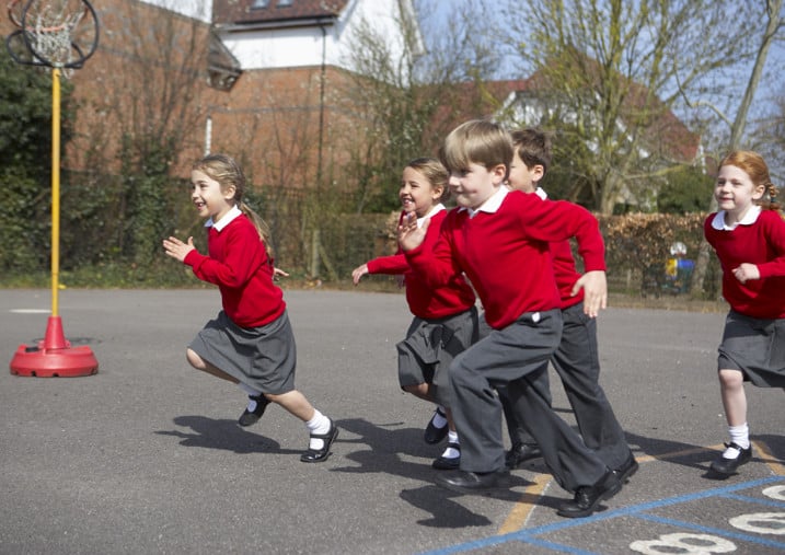 Smiling children in red school uniform running together across sunny tarmac, with bare trees and houses behind
