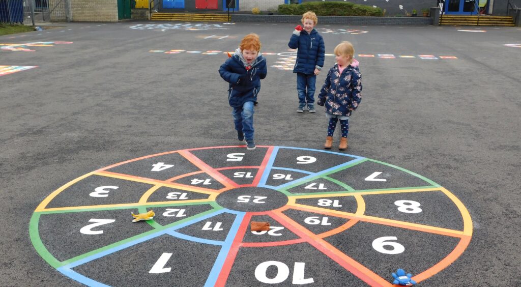 Three young children heading towards a rainbow number target marking, 1 to 20 with 25 in the middle, with soft toys on it
