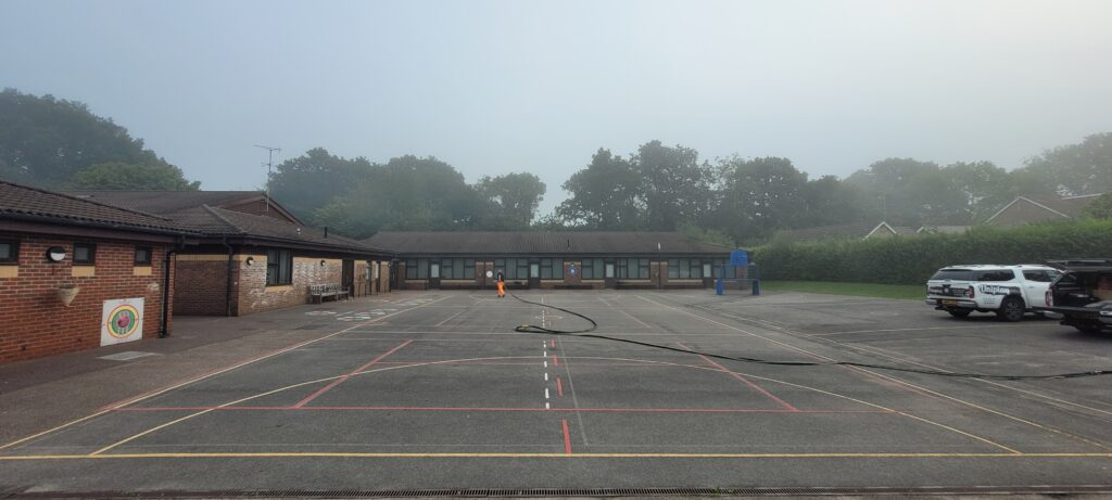 Faded court line markings on a misty school playground, with a Uniplay pickup parked and a worker in hi-vis running a long hose