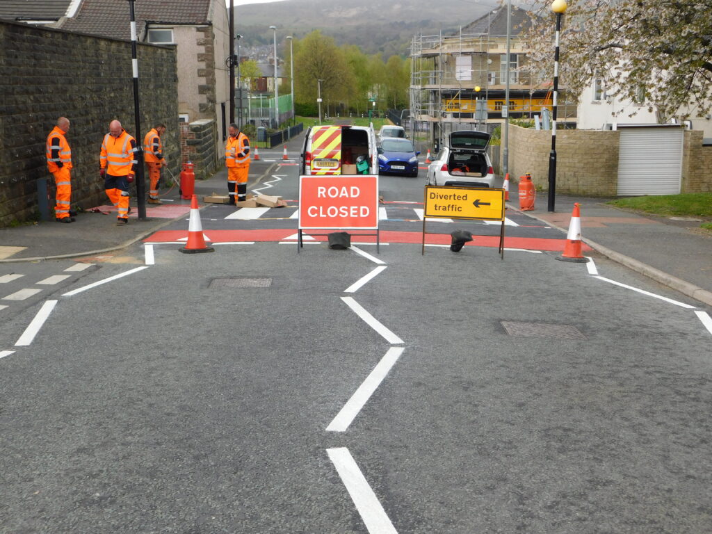 Road marking crew in hi-vis behind Road Closed and Diverted traffic signs, with fresh white zigzags and red surfacing
