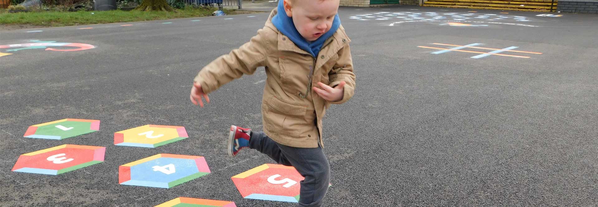 Young boy in a beige coat hopping along a numbered hexagon trail marking, with a noughts and crosses grid on the tarmac behind