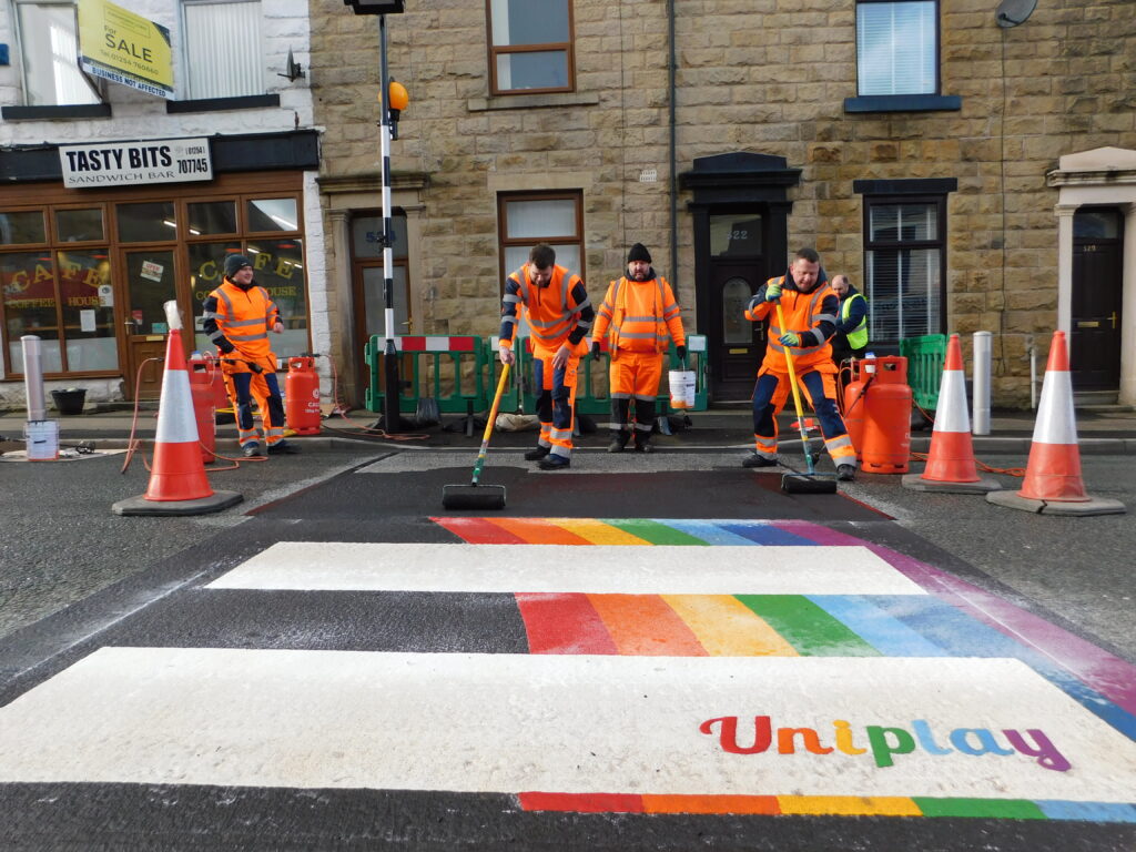 Road marking crew in orange hi-vis working on a rainbow-edged zebra crossing with Uniplay lettering on a town street