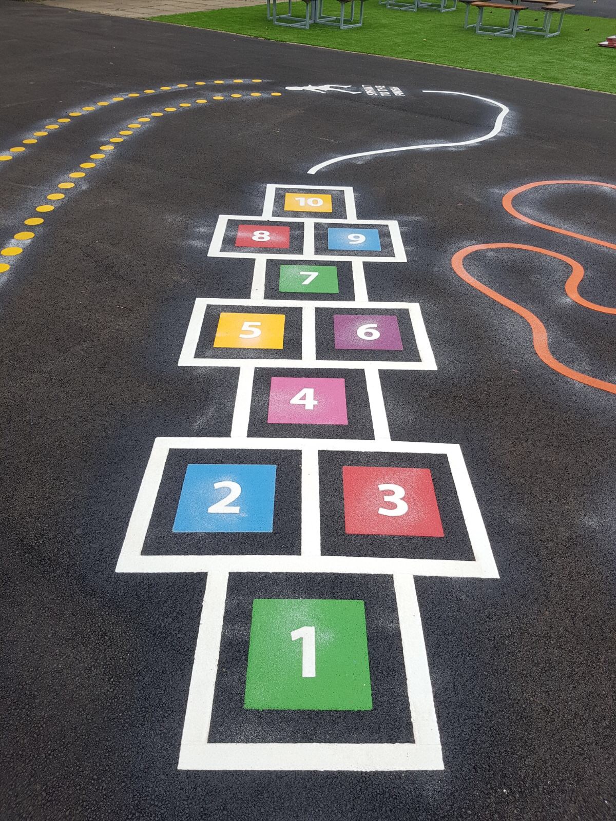 Hopscotch numbered 1 to 10 in coloured squares with white outlines on new tarmac, beside dotted and wiggly trail markings
