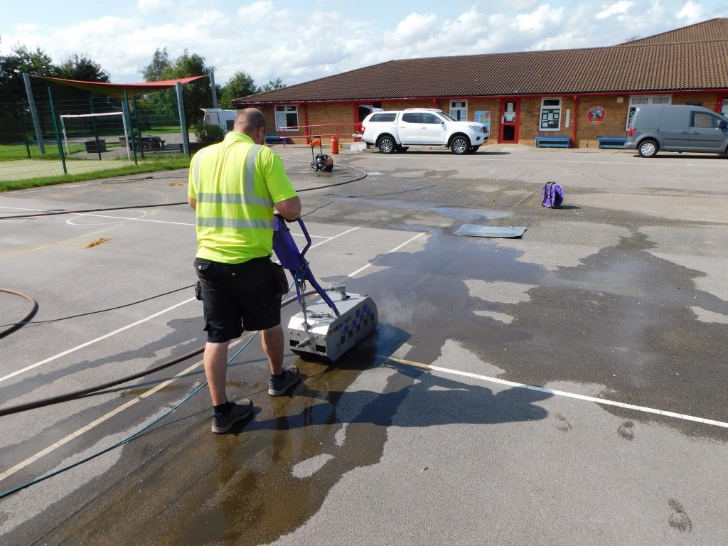 Operative in yellow hi-vis removing old white court line markings from a school playground with a water jetting machine