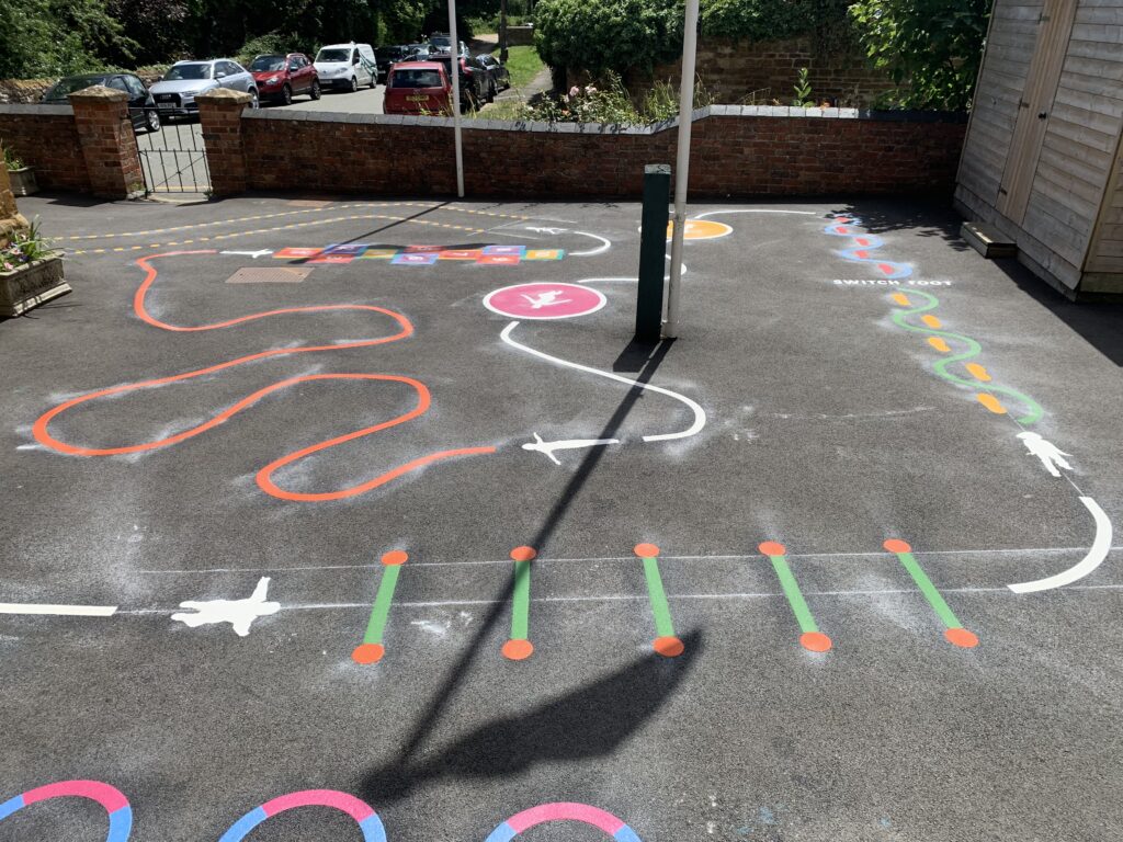 Small walled school playground with orange wiggle line, white aeroplane trail, switch foot hopping path and jump bar markings