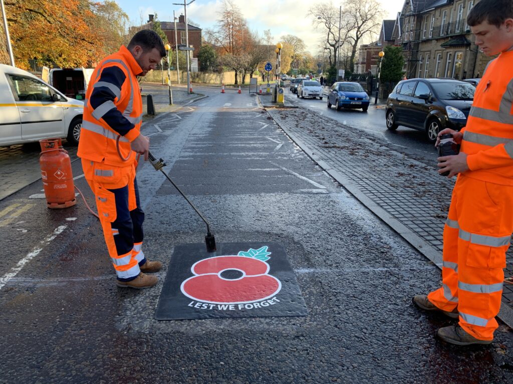 Installer heating a preformed thermoplastic poppy marking reading Lest We Forget with a gas torch on an autumn street
