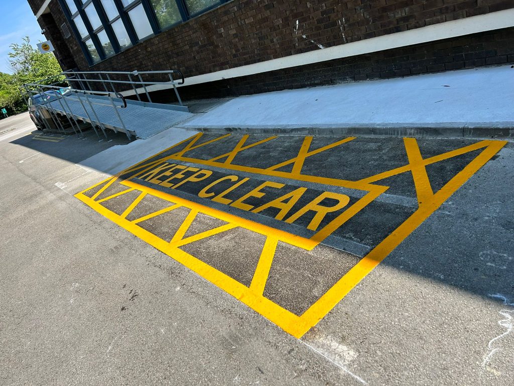 Yellow Keep Clear box marking with hatched edges at the foot of an access ramp beside a brick building