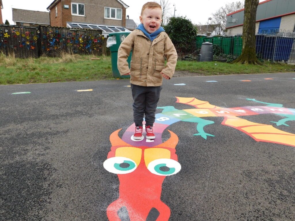 Laughing toddler in red trainers standing on a thermoplastic dragon hopscotch with a numbered body and green legs on a path