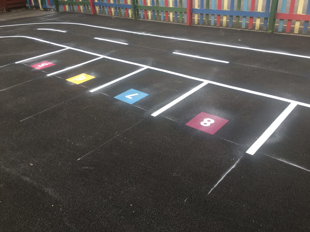 White-lined play road marking with numbered parking bays in pink, yellow, blue and maroon on new tarmac by a multicoloured fence