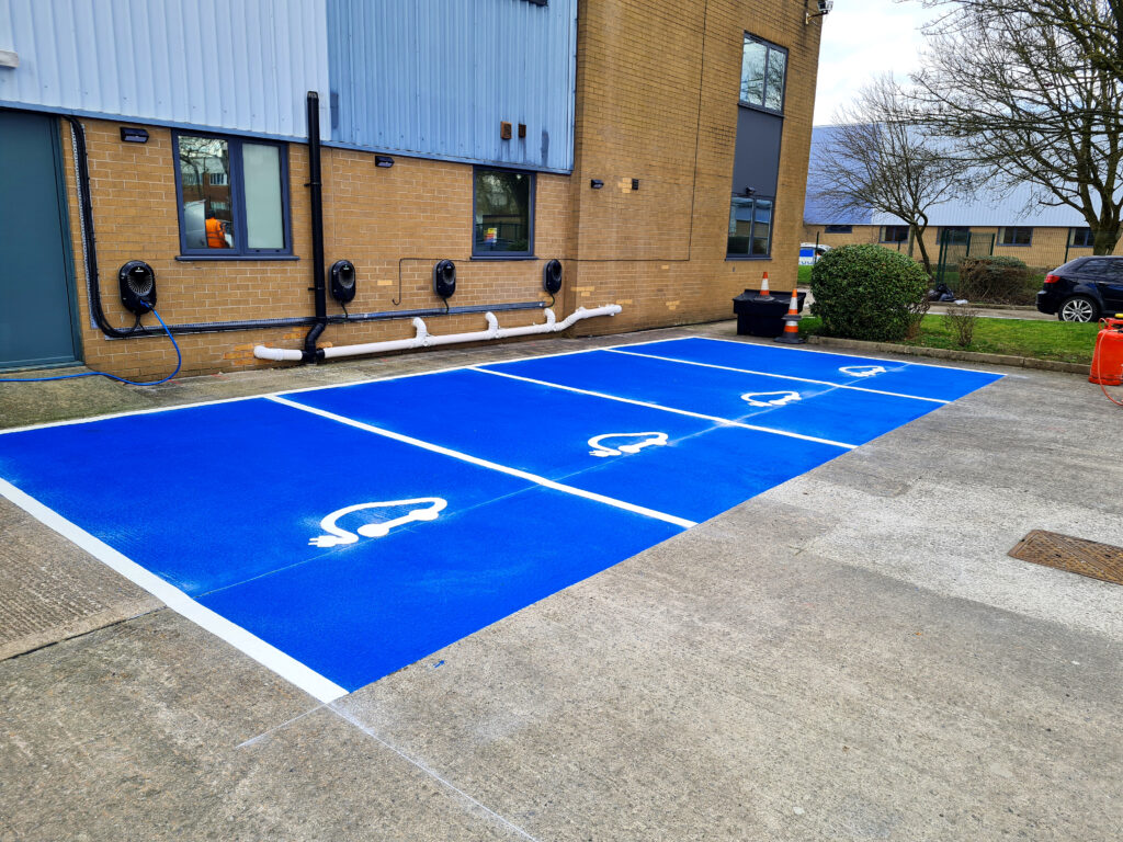 Four blue thermoplastic EV charging bay markings with white electric car symbols beside wall chargers outside a brick business unit