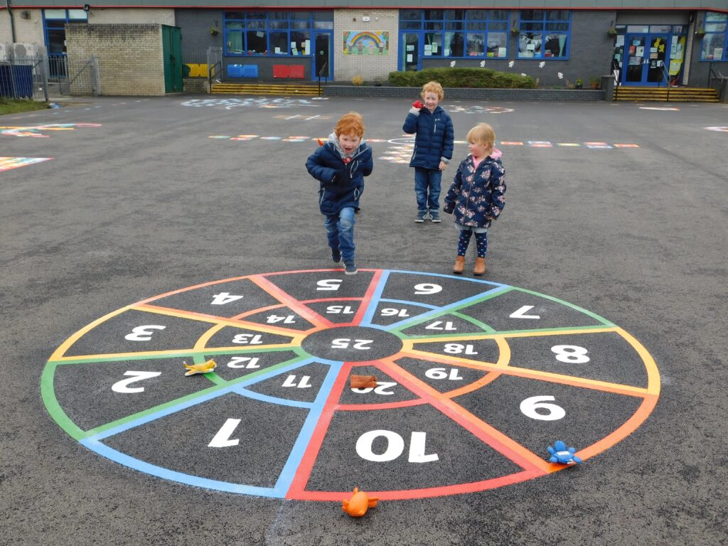 Three young children by a rainbow-ringed number target marking with soft toys thrown onto it, in front of a school building