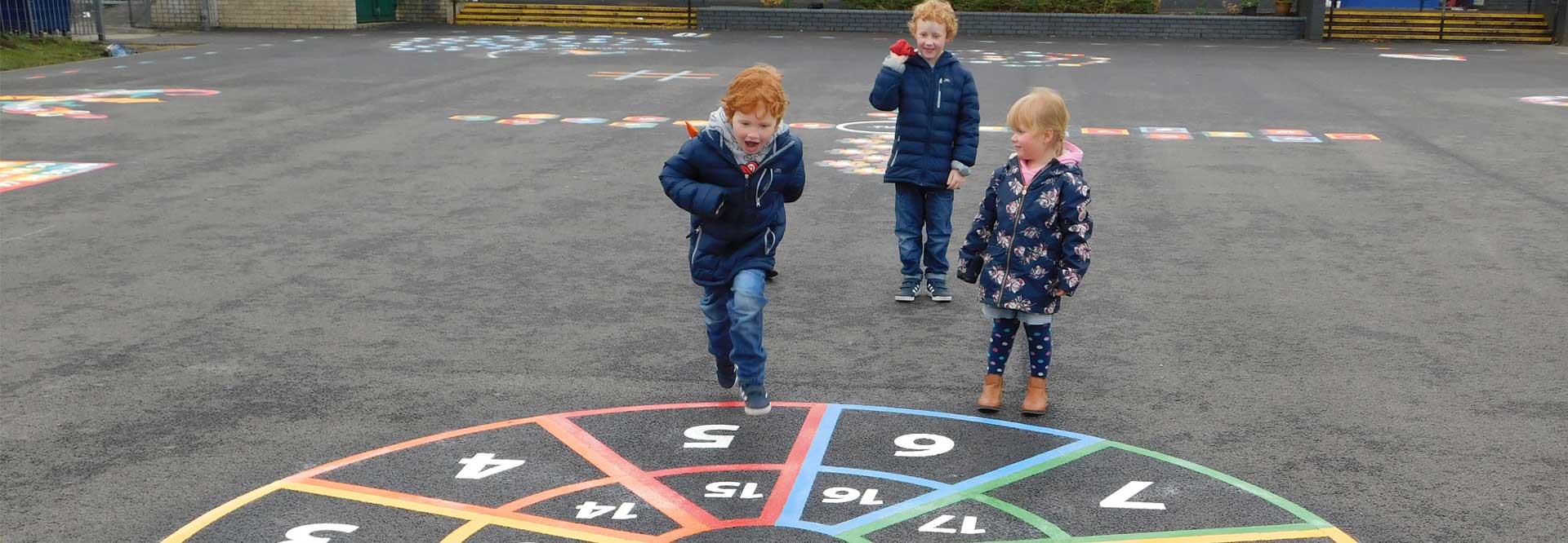 Wide view of a boy leaping towards a numbered target circle as two children watch on a playground dotted with markings