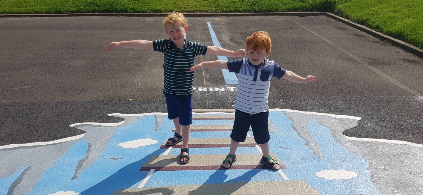 Two young boys with arms outstretched balancing on a 3D bridge illusion painted over sky and cliffs on playground tarmac