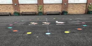 Duck duck goose marking with a ring of coloured spots around duck and goose figures, by benches on a school playground