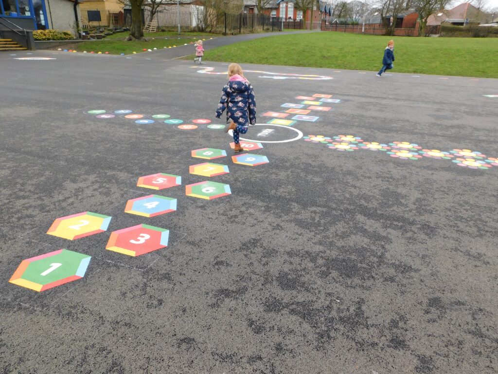 Young girl hopping along a numbered hexagon hopscotch on a school playground, with flower and trail markings beyond