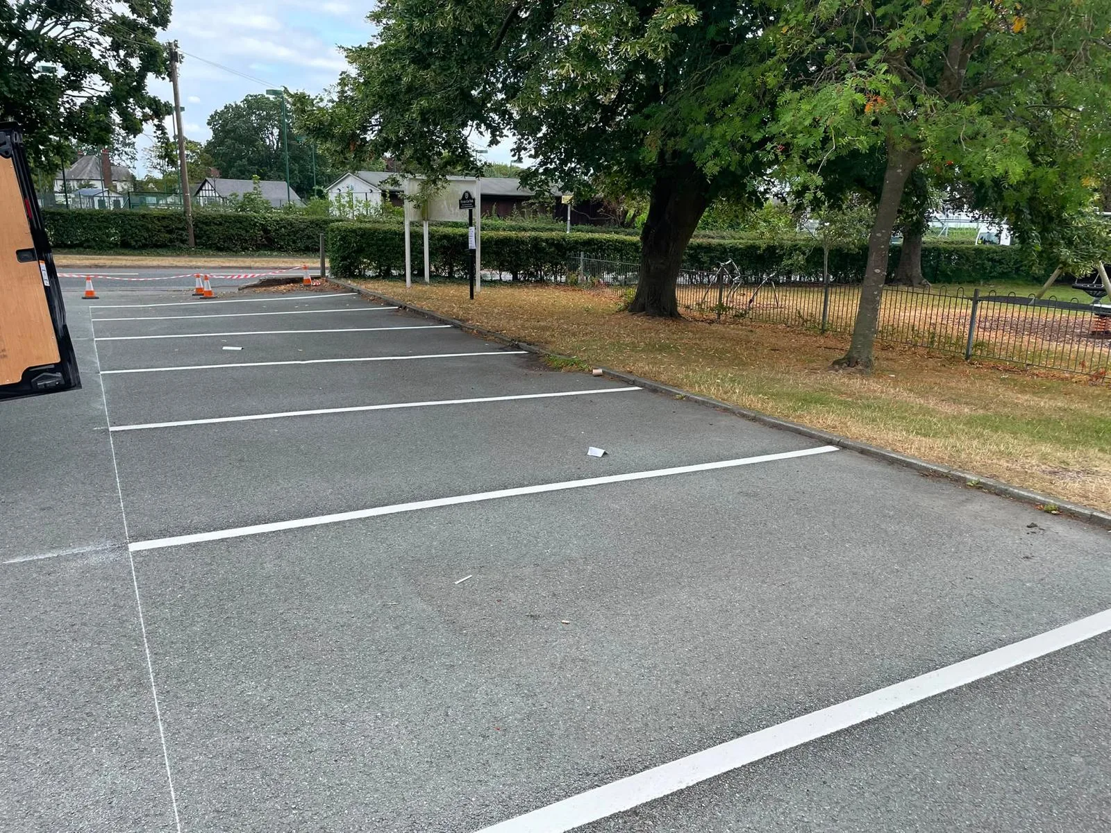 New white parking bay line markings in a tree-lined car park beside a fenced play area, with cones and tape across the entrance
