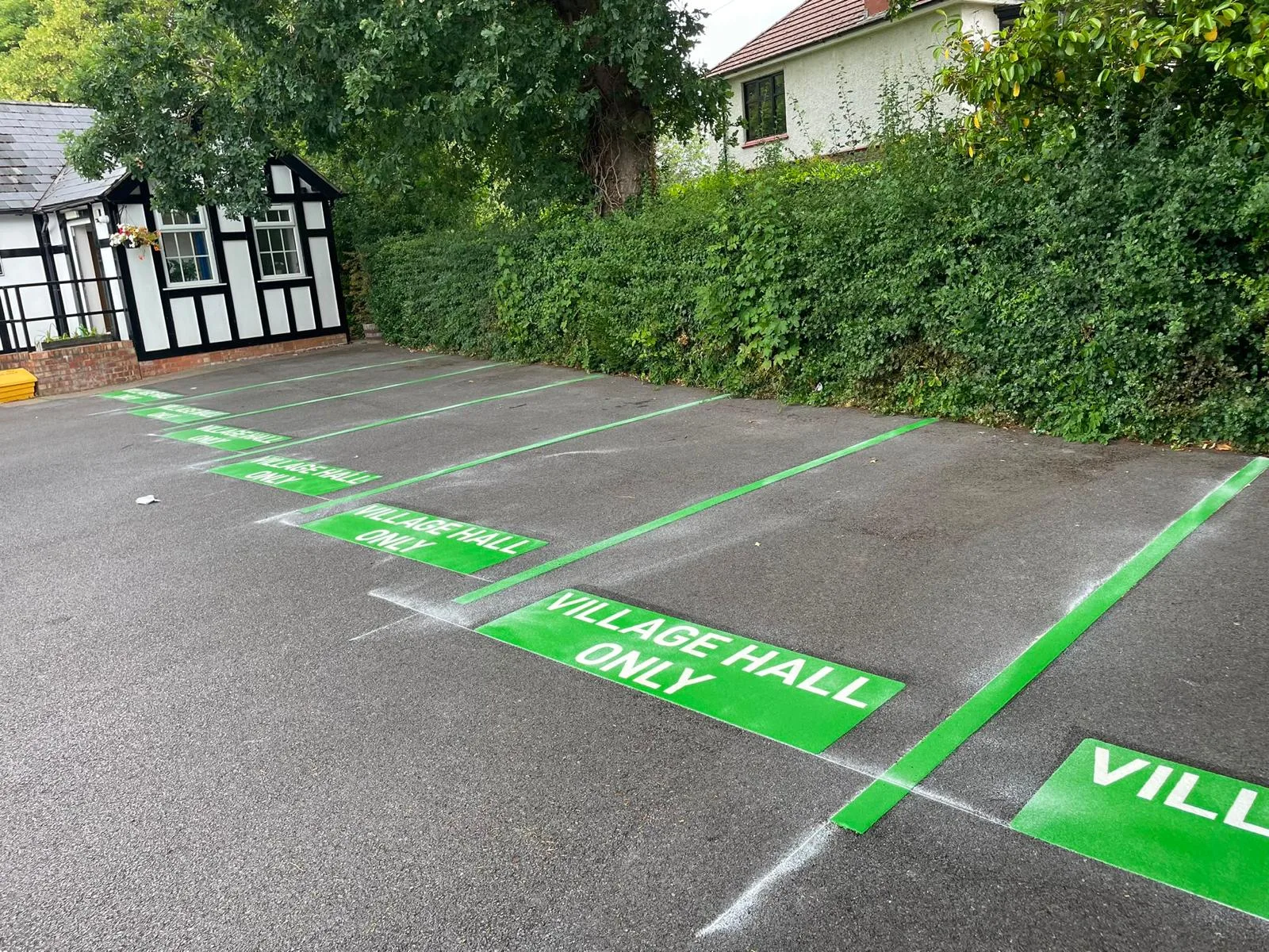 Row of green VILLAGE HALL ONLY bay markings along a tall hedge, leading to a black and white timber-framed building