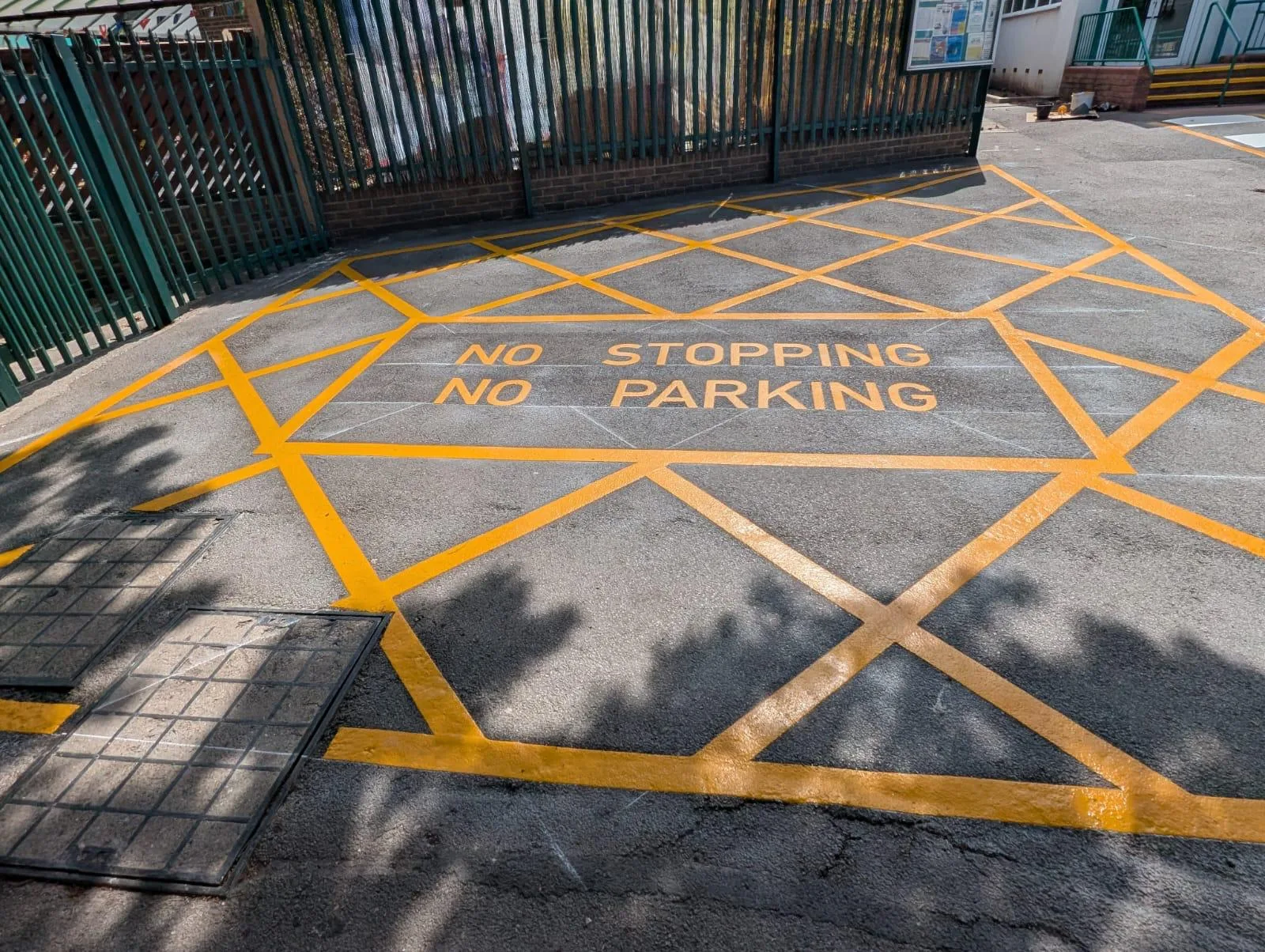Front-on view of a yellow hatched NO STOPPING NO PARKING zone beside green palisade railings, with two inspection covers