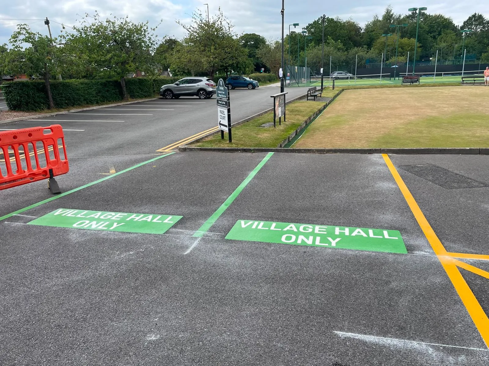Two green VILLAGE HALL ONLY bay markings and a yellow line beside a bowling green, with tennis courts in the distance