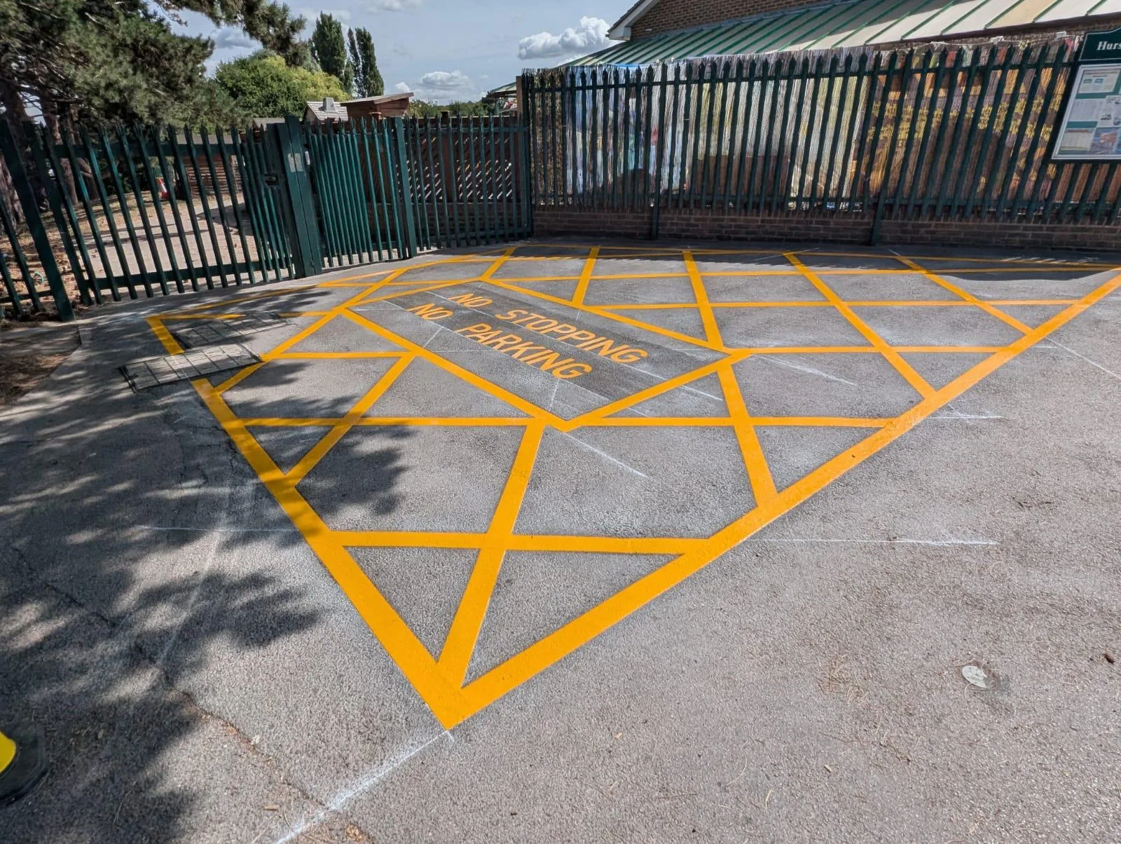 Wide-angle view of yellow cross-hatching and NO STOPPING NO PARKING road markings by green palisade gates and a noticeboard