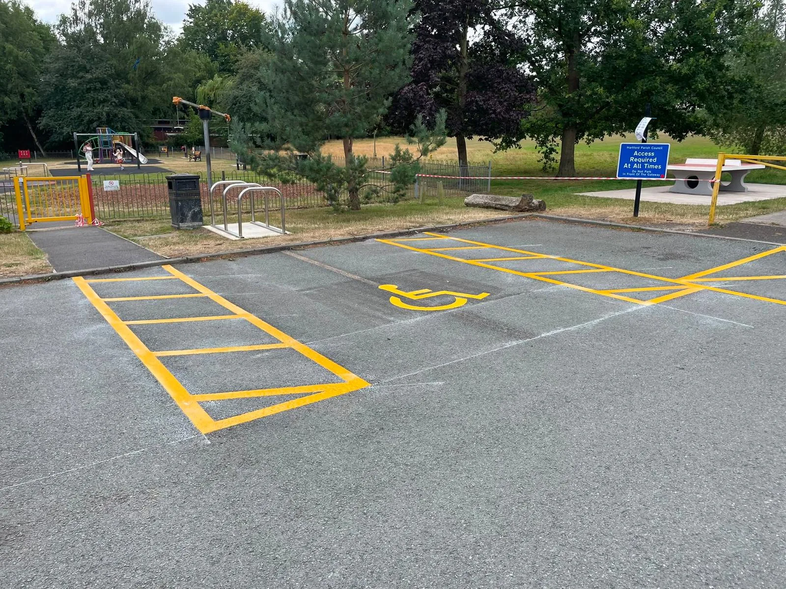 Yellow disabled bay marking with a wheelchair symbol between hatched access zones, next to a park play area and cycle stands