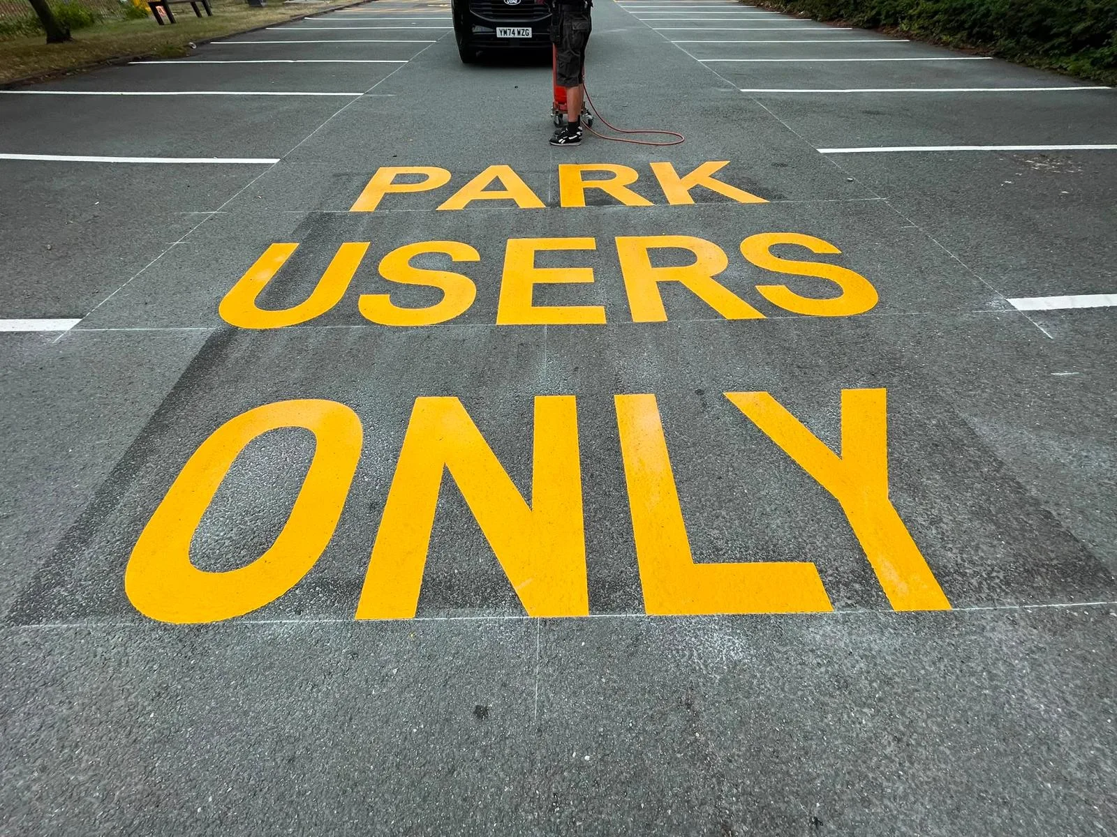 Large yellow PARK USERS ONLY thermoplastic lettering in a car park aisle, with an installer and gas bottle trolley behind