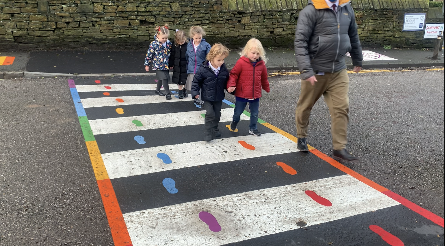 Pupils on a rainbow-edged crossing marking with coloured footprints outside Turton and Edgworth CE Methodist Primary School