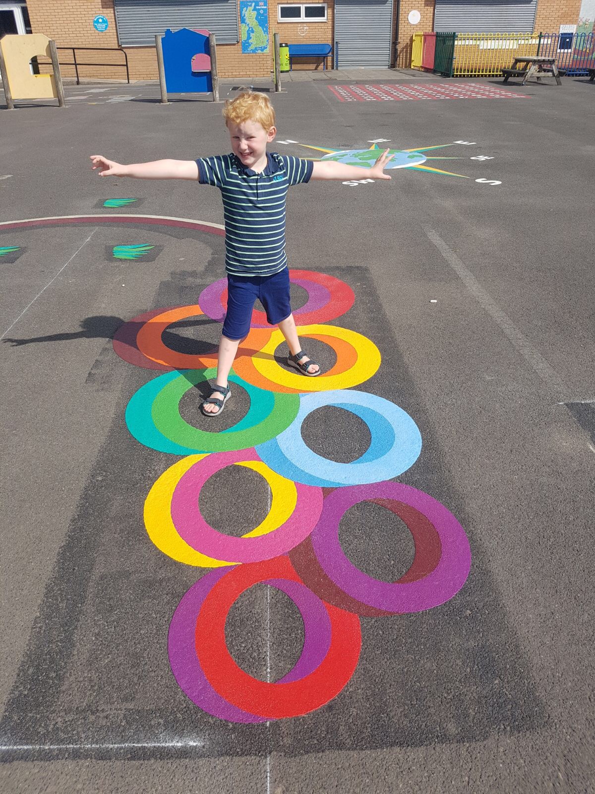 Boy with arms outstretched on 3D-effect rainbow ring markings, with a compass and red number grid on the playground behind
