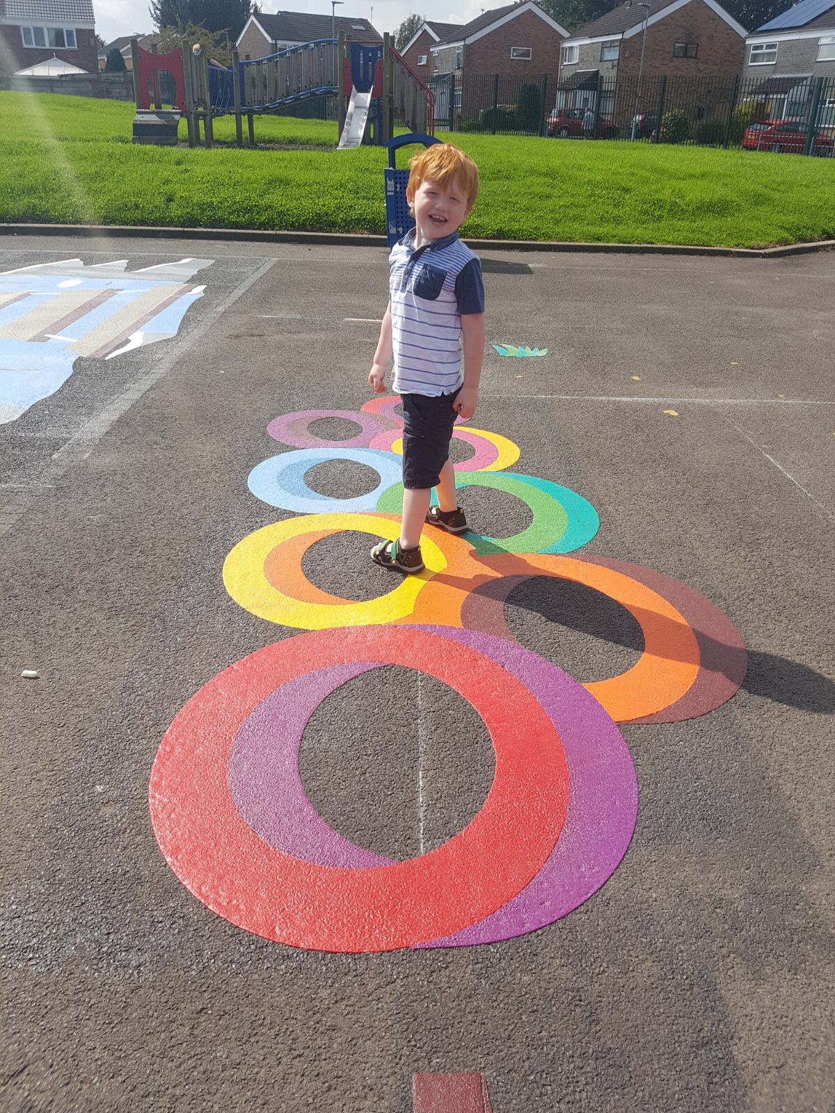 Smiling boy standing on overlapping 3D-effect rainbow rings, with a climbing frame, slide and houses behind