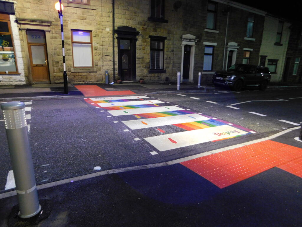 Zebra crossing marking with rainbow stripes, coloured footprints and Uniplay lettering on a terraced street, lit at night