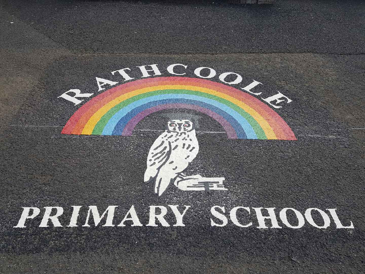 Rathcoole Primary School logo laid in thermoplastic on tarmac, with a rainbow arching over a white owl perched on books