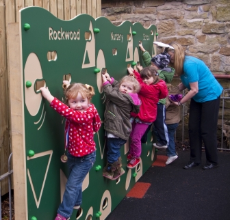 Nursery children climb a green Rockwood Nursery climbing wall with round holds and shape cut-outs as an adult looks on