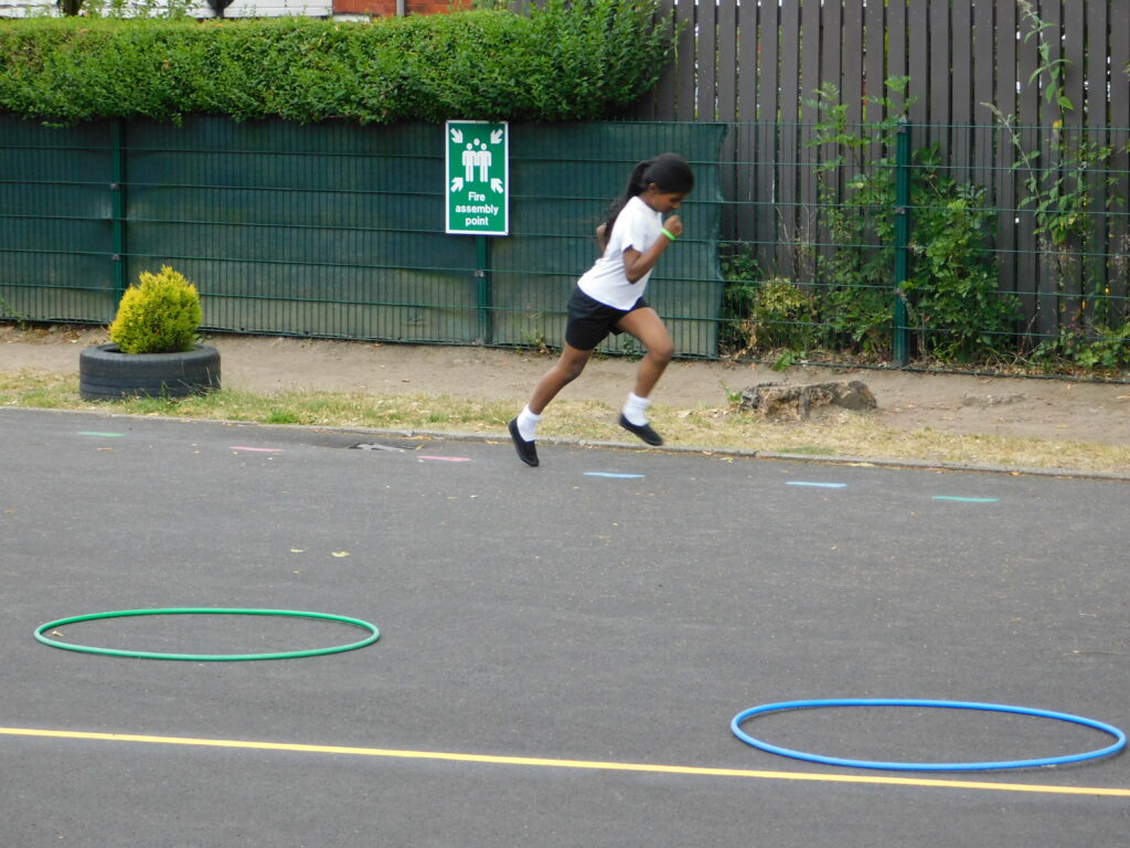 Roe Lee School pupil in PE kit leaps across the playground between green and blue hoops beside a yellow line marking