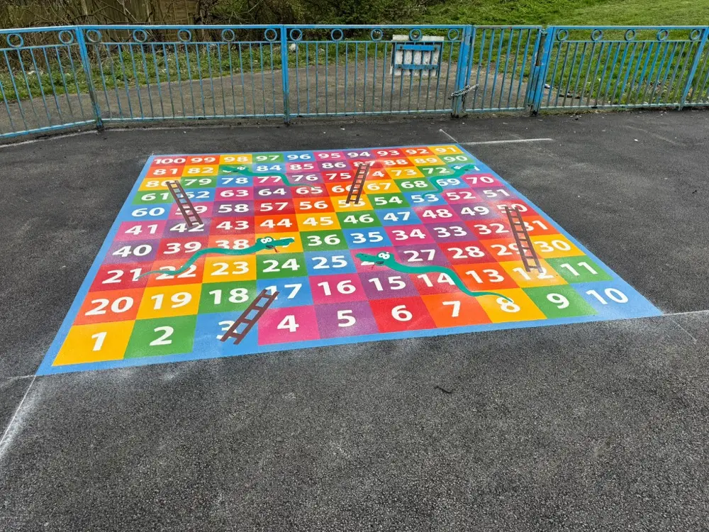 1 to 100 Snakes and Ladders board viewed square-on beside a blue railing fence and grass bank