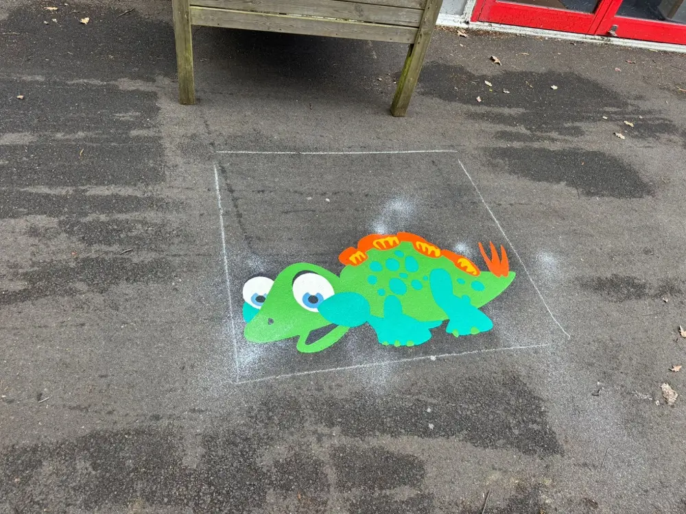 Green and orange stegosaurus dinosaur thermoplastic marking on a damp tarmac playground beside a wooden bench