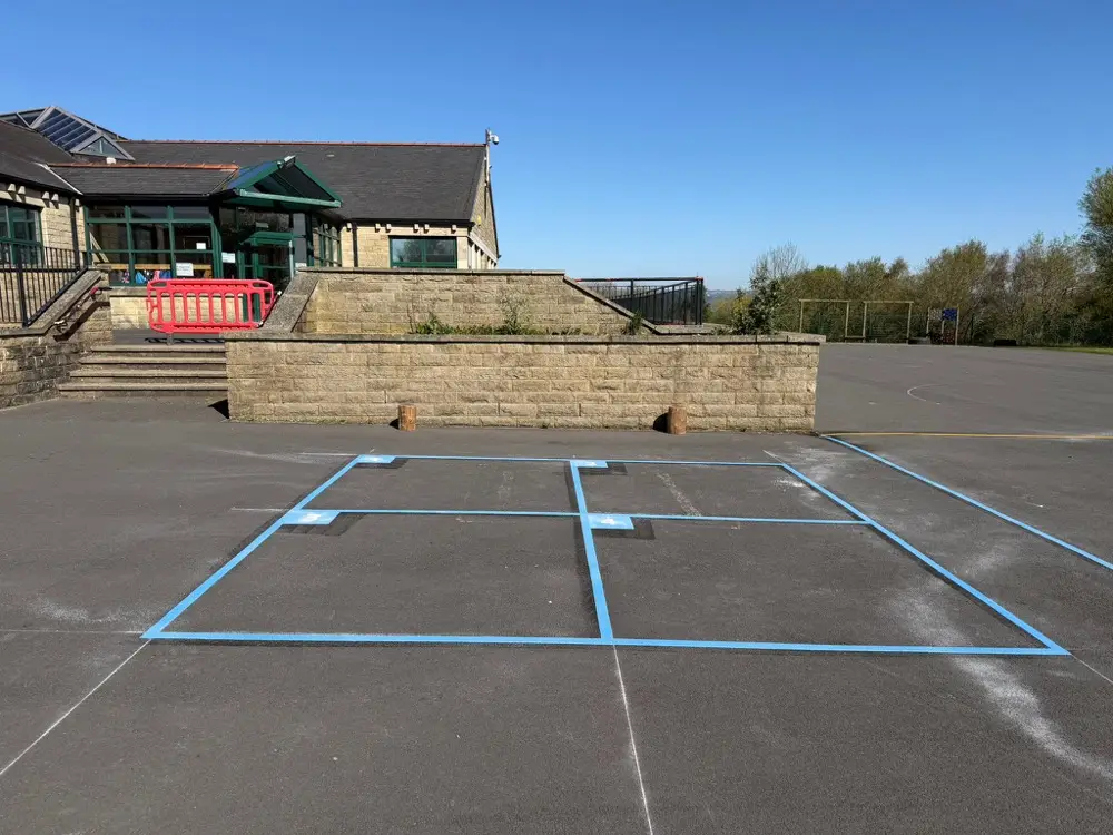Pale blue Four Square Game outline on worn tarmac under a clear sky beside a stone retaining wall