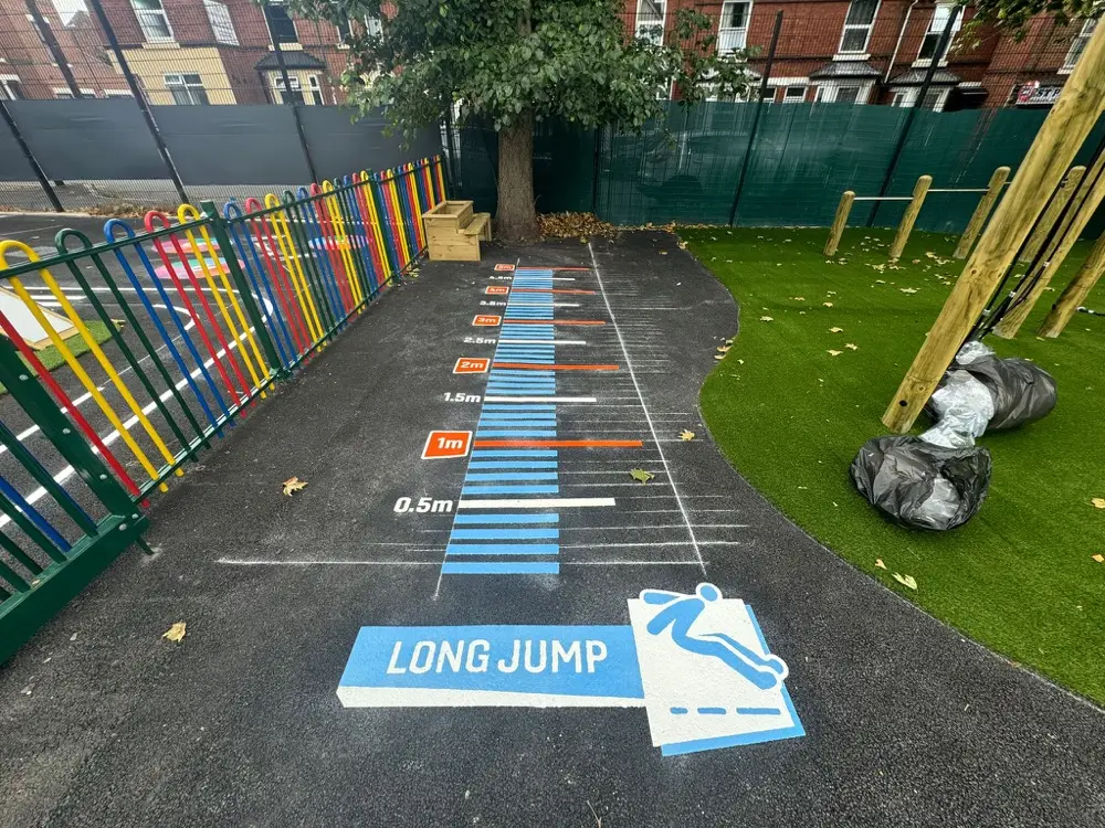 Blue Long Jump track with metre markers and a take-off graphic on tarmac beside a rainbow railing and artificial grass