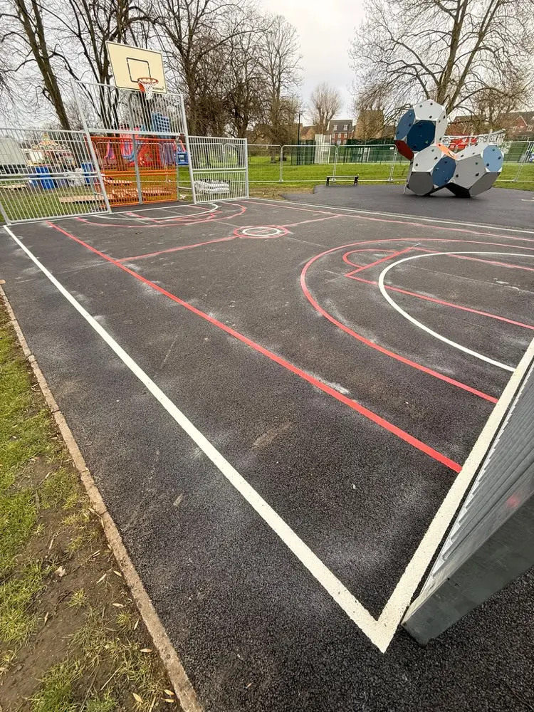 Multi-sport court markings in red and white lines on dark tarmac beside a basketball hoop and climbing frame