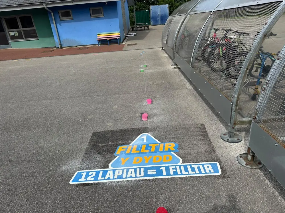 Welsh Filltir y Dydd Mile a Day marker reading 12 lapiau = 1 filltir, beside a covered bike shelter on school tarmac
