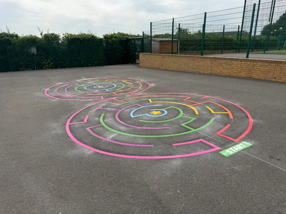 Double circular maze markings photographed from a low angle on tarmac beside a brick wall and hedge