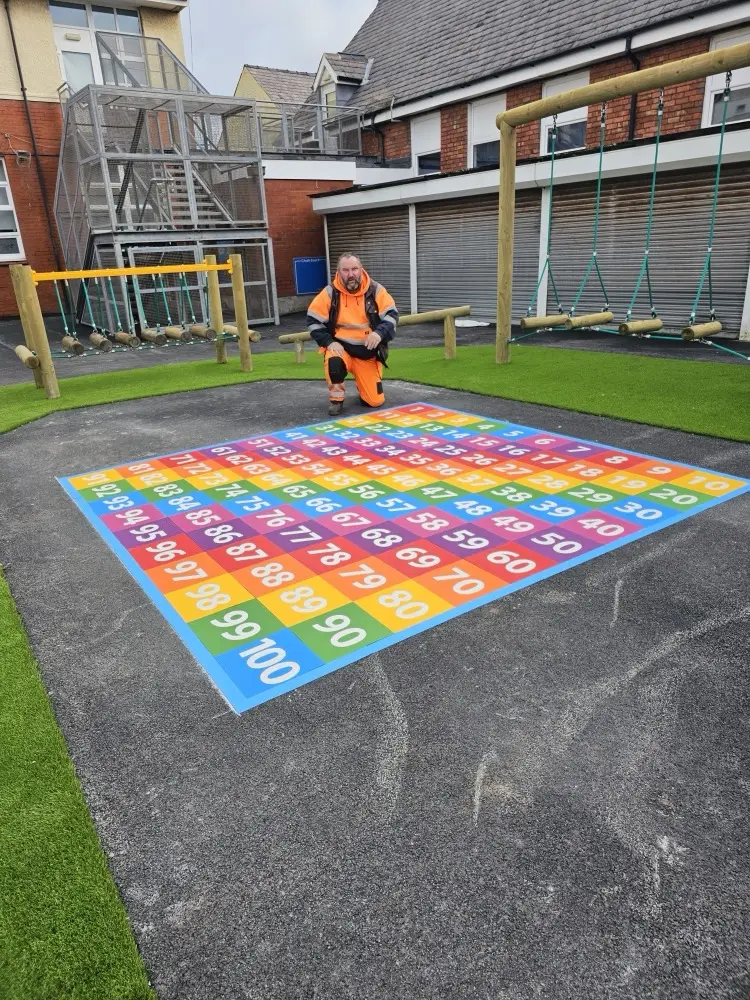 Installer in hi-vis behind a newly laid rainbow thermoplastic 1 to 100 number grid, between a rope swing frame and climbing net